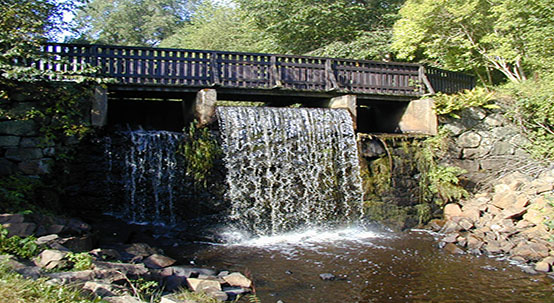 Bilden visar en gångbro och under bron forsar det vatten. Runt omkring syns gröna träd.