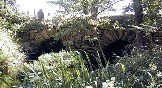 Bilden visar en stenmur som har två stycken valv där det rinner igenom vatten. Mycket grönska runt omkring.